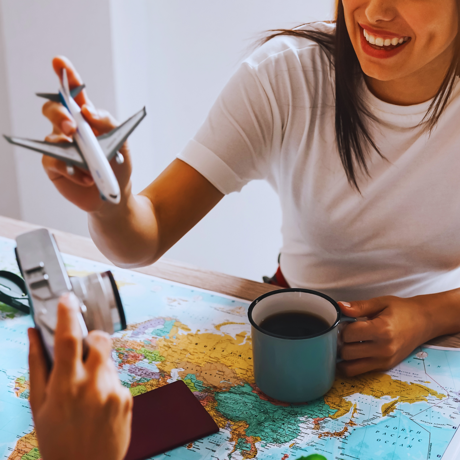 Smiling traveler sipping from a teal mug while a friend holds a toy airplane over a world map—capturing the spirit of unique gifts for globetrotters.