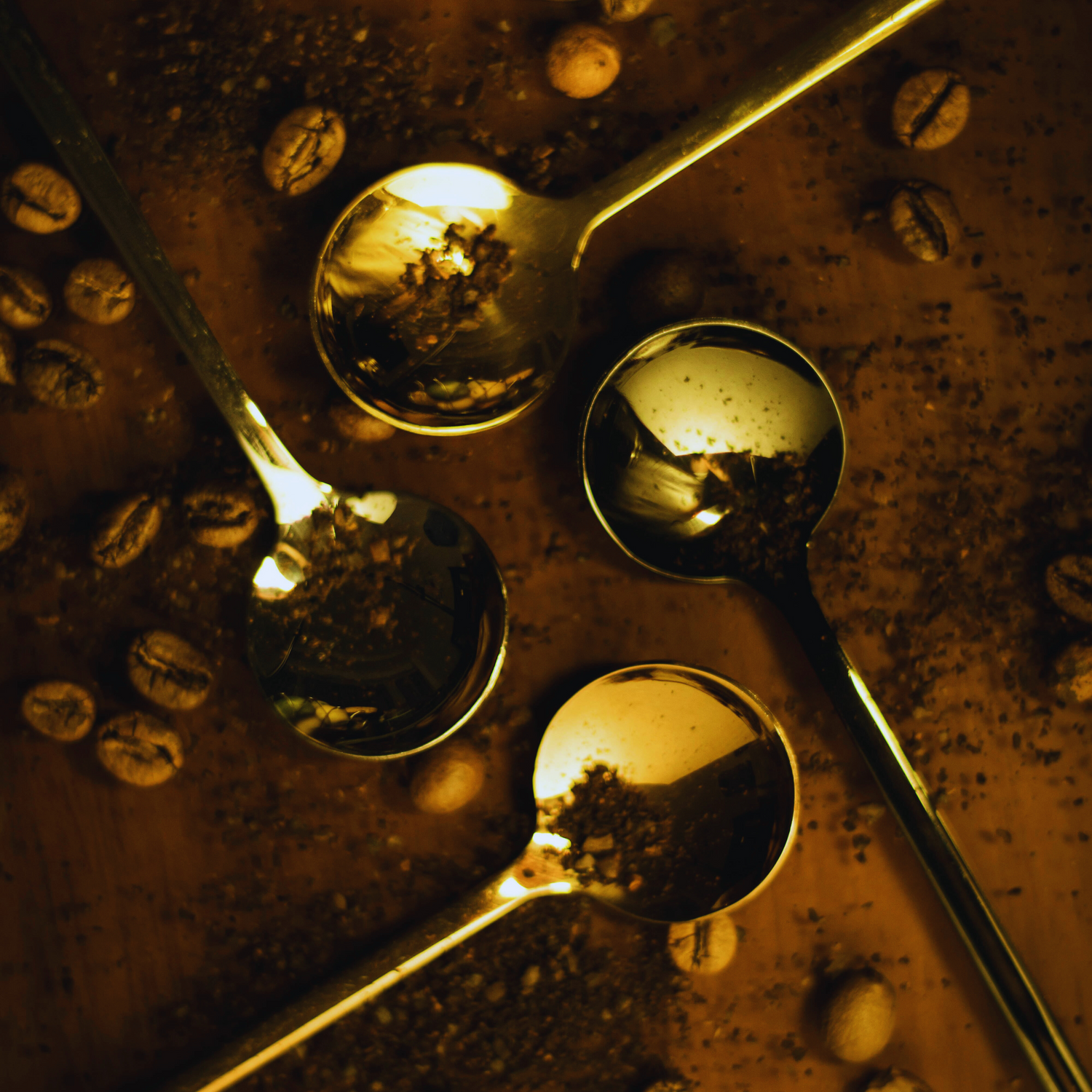 Flat-lay of four reflective gold coffee spoons resting on scattered coffee beans and grounds, illustrating unique gifts for passionate coffee lovers.