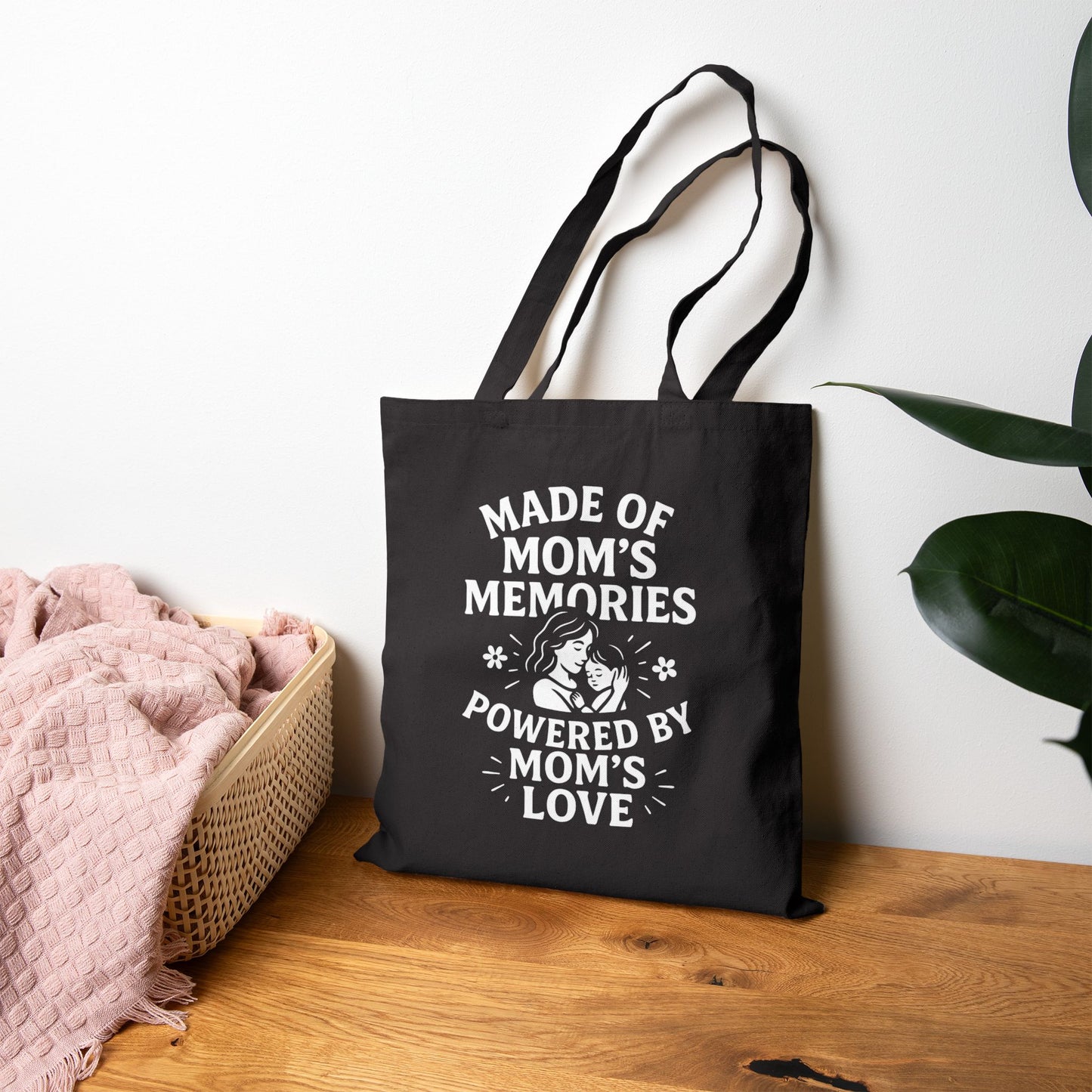 Black tote bag displaying heartfelt mom‑memory design, propped against wall on wooden surface with greenery.