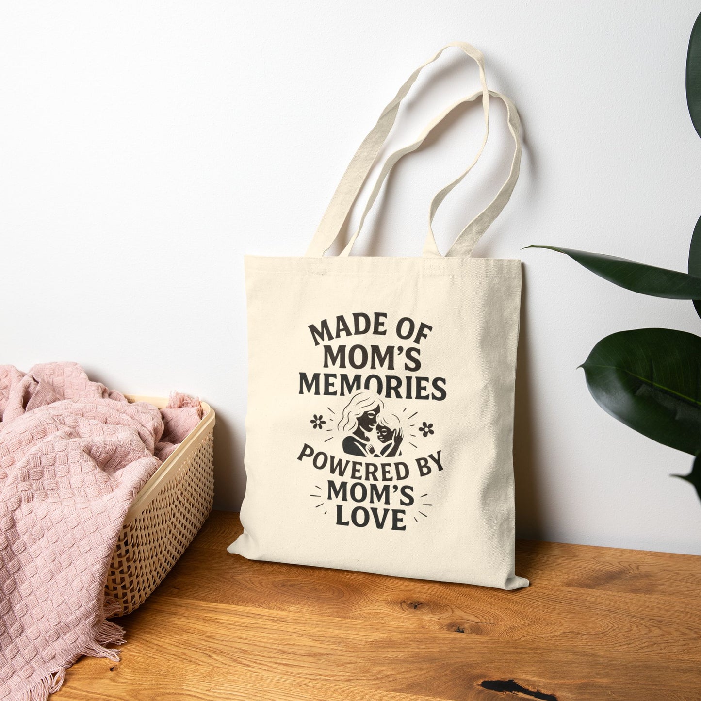Beige cotton tote bag with sentimental mom quote hanging on wall above wooden table and cozy blanket.