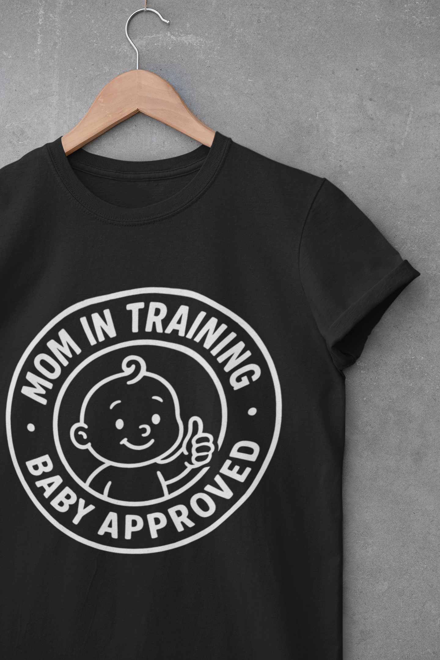 Black funny mom shirts design with “Mom in Training • Baby Approved” graphic displayed on hanger against gray wall.