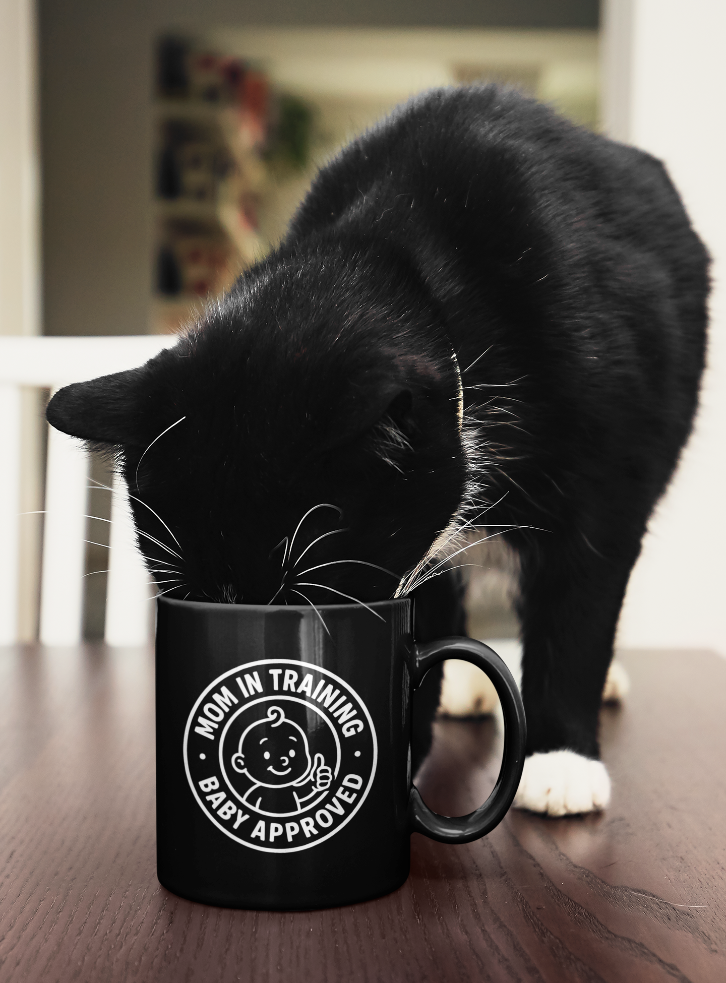 Black ceramic mug with “Mom in Training, Baby Approved” design being sniffed by a curious black-and-white cat on a wooden table.