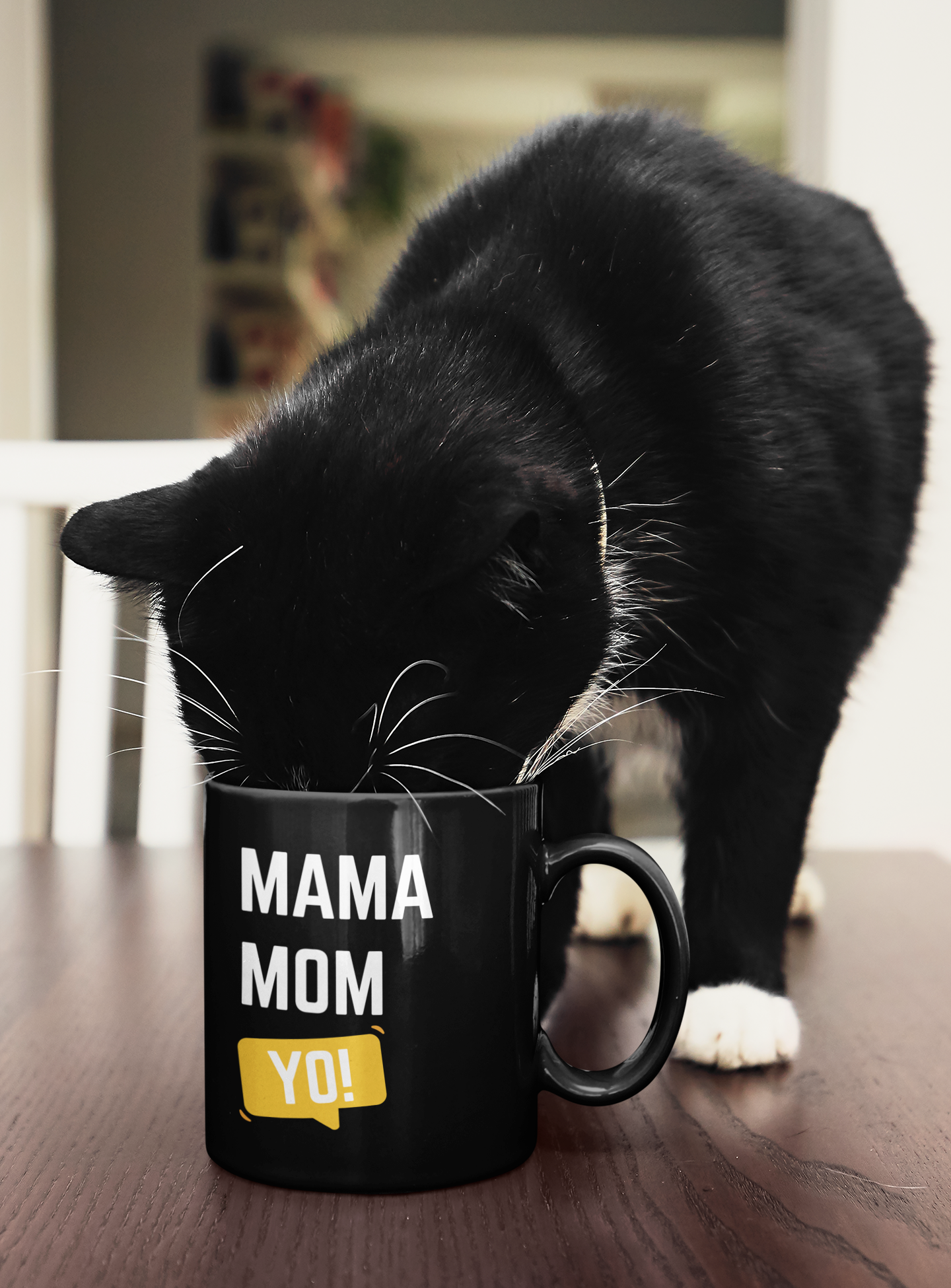 Black ceramic mug with “Mama Mom Yo!” design being curiously sniffed by a black-and-white cat, adding playful charm to the morning coffee vibe.