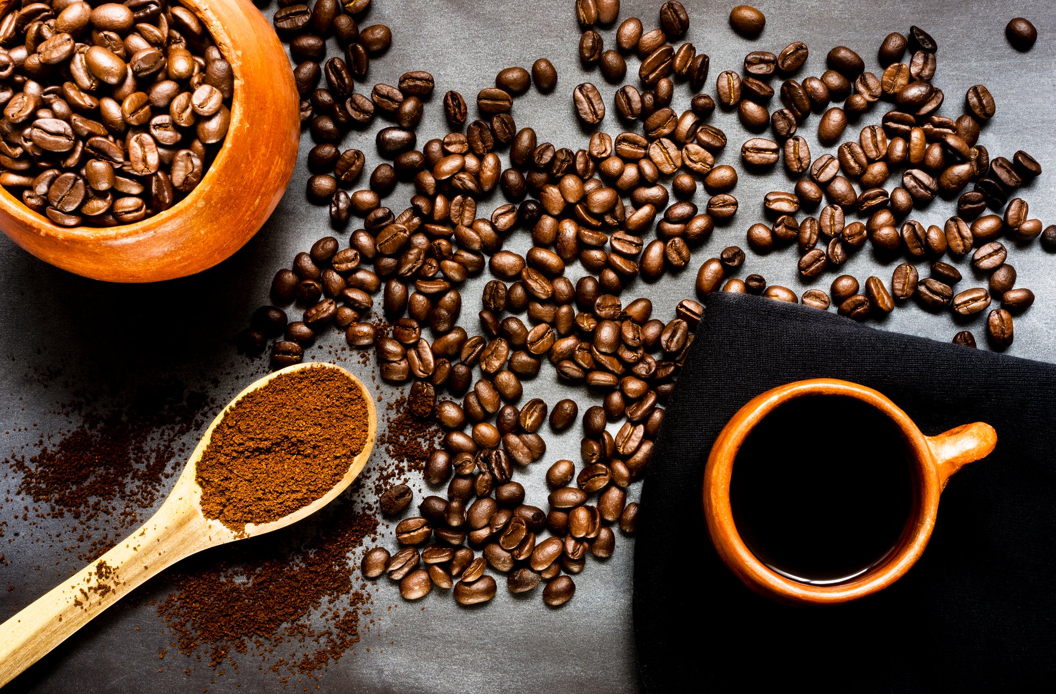 Flat-lay of glossy roasted coffee beans, fragrant grounds in a wooden scoop, and a rustic clay cup of black coffee on slate—rich, sensory scene perfect for coffee-lover unique gifts imagery.