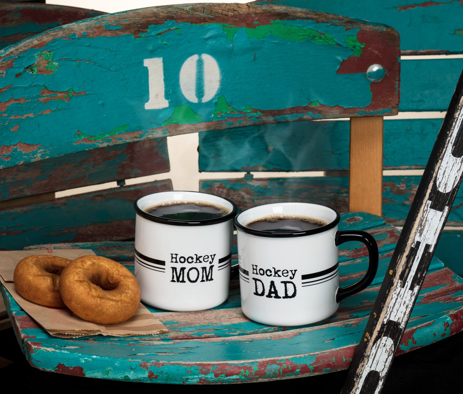 Hockey MOM and Hockey DAD enamel mugs filled with hot coffee, set beside donuts and a vintage hockey stick on a rustic bench.