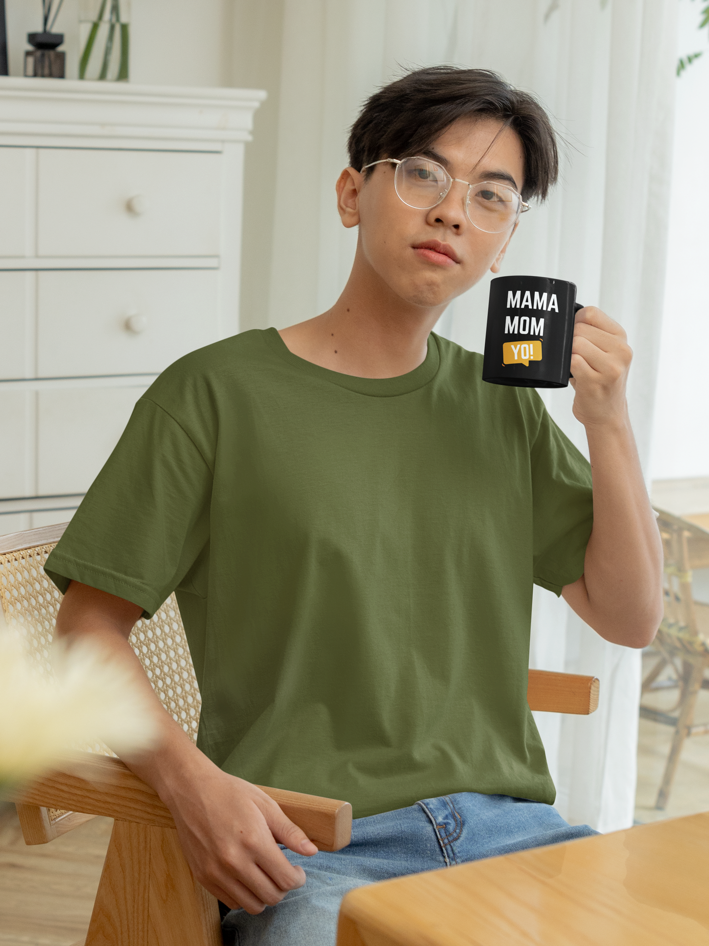 Young man sitting at a kitchen table holding a black “Mama Mom Yo!” ceramic mugs design, perfect for a funny Mother’s Day gift.