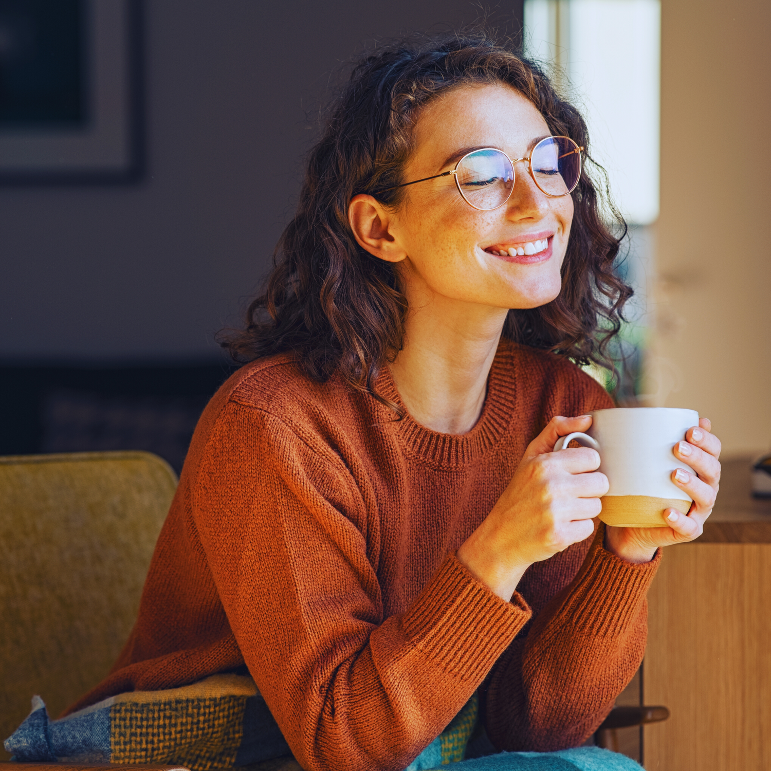 Smiling woman in cozy sweater enjoying morning coffee from ceramic mugs near bright window