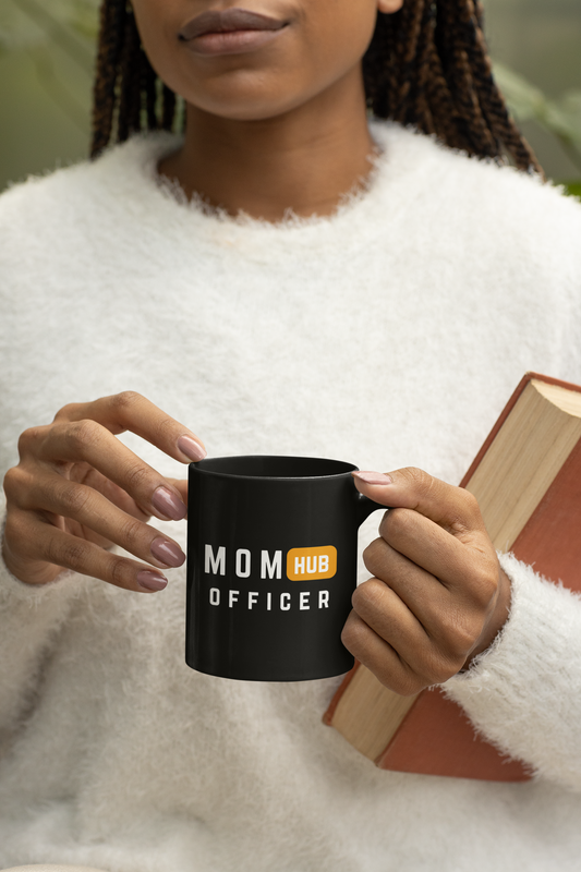 Black ceramic mug with bold “Mom Hub Officer” text held by woman in soft white sweater, ideal gift for multitasking moms.