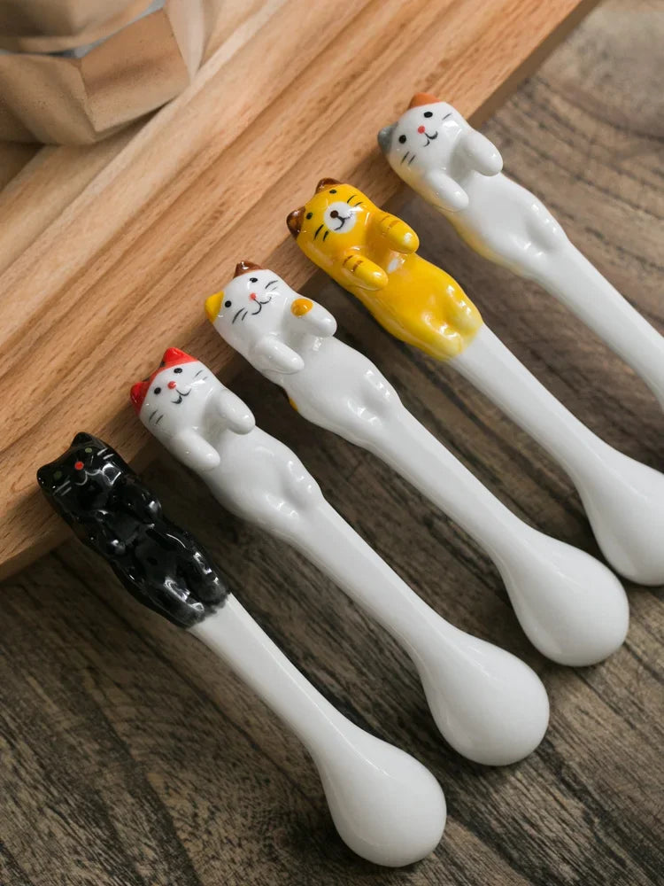 Line‑up of five assorted ceramic cat coffee spoons on wooden board—colorful set of collectible funny gifts for tea or espresso.








