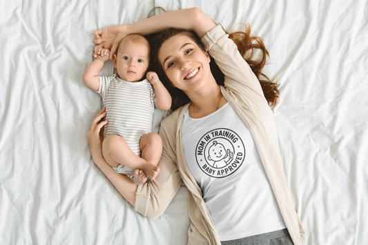 Smiling mom wearing white “Mom in Training • Baby Approved” tee cuddling newborn on bed, showcasing comfy mom shirts for new mothers.