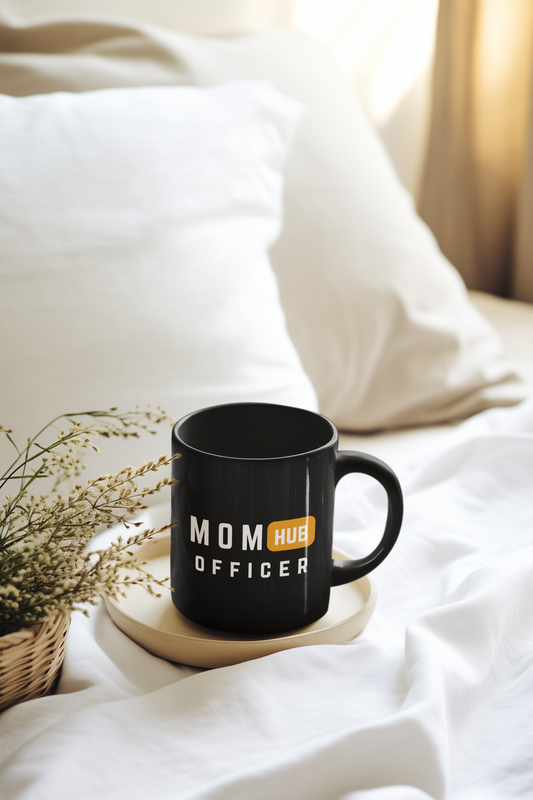 Black ceramic mug with “Mom Hub Officer” design placed on a cozy bed tray, perfect for moms who handle every mission before coffee.