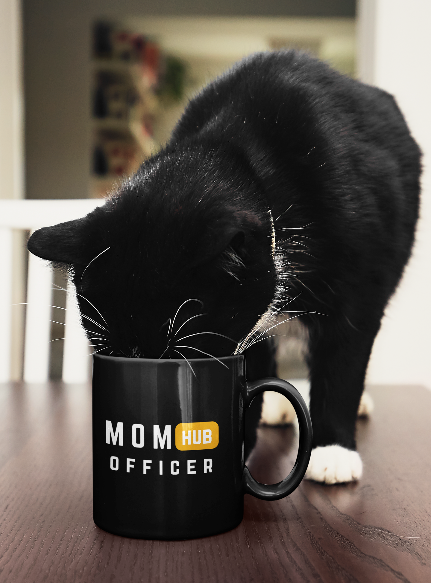 Black ceramic mug with “Mom Hub Officer” design being sniffed by a curious black-and-white cat on a table, adding a playful lifestyle touch.