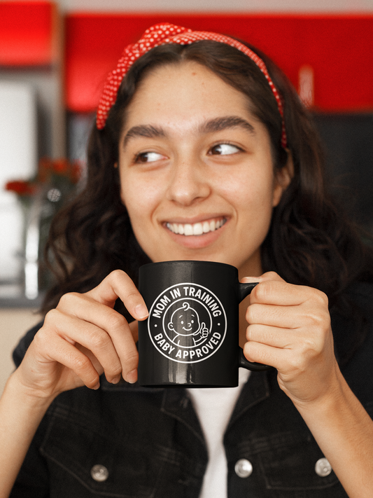 Woman smiling and holding black ceramic mug with “Mom in Training, Baby Approved” design, perfect gift for new or expecting moms.