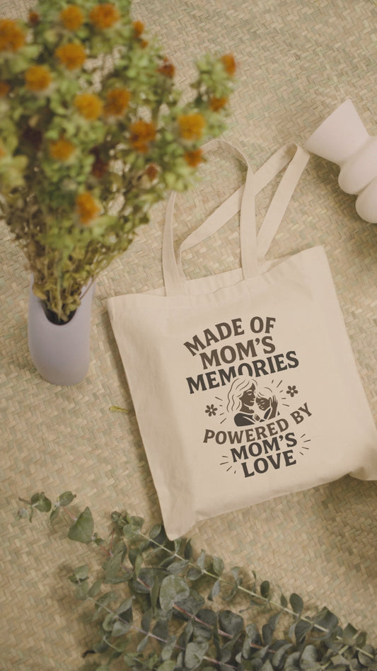 Beige cotton tote bag with “Made of Mom’s Memories Powered by Mom’s Love” print, styled on woven mat beside flowers.